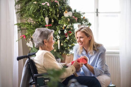 Blonde woman and older lady in wheelchair exchange a gift in front of a Christmas tree